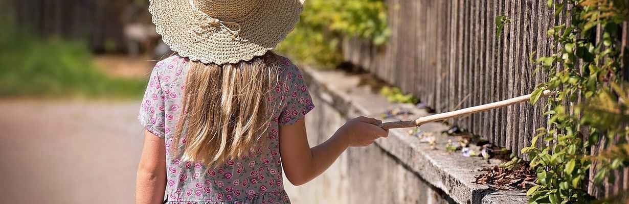 Girl wearing wide brim straw hat drags a stick along picket fence